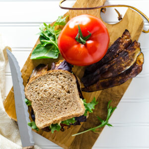 Vegan Gluten Free BLT sandwich on a cutting board with a tomato and strips of eggplant bacon