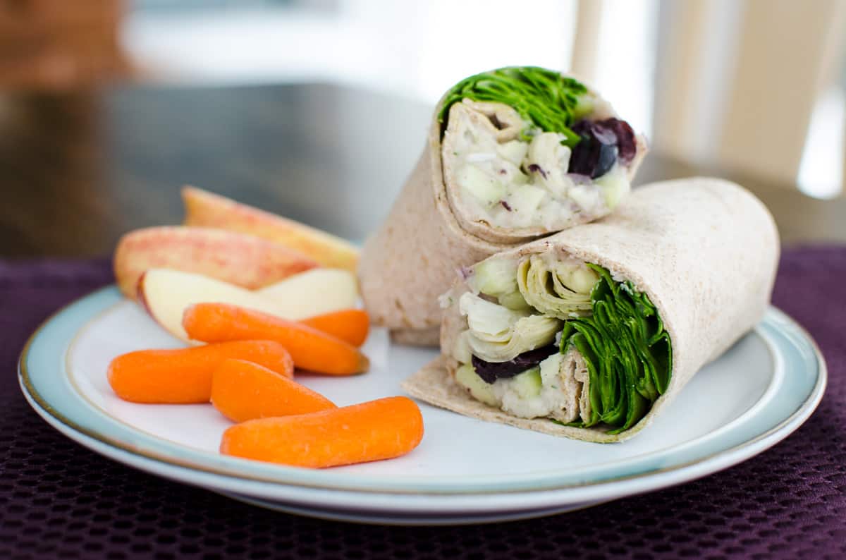 Vegan greek sandwich wrap cut in half, on a plate with carrots and sliced apple