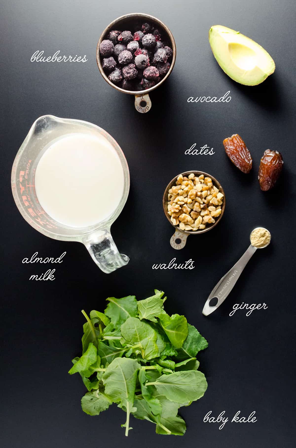Overhead view of super smoothie ingredients, including blueberries, avocado, dates, walnuts, ginger, baby kale, and almond milk