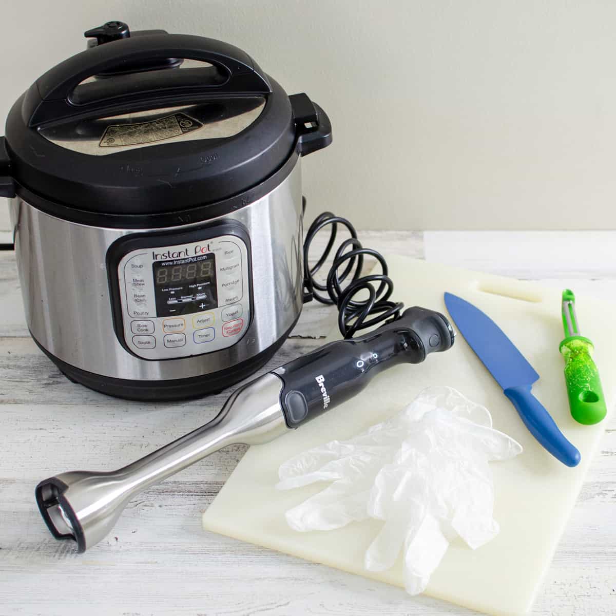 An Instant Pot, stick blender, chef knife, vegetable peeler, food prep gloves, and a cutting board