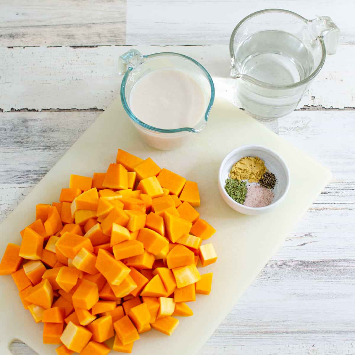 chopped butternut squash on a cutting board next to measuring cups filled with plant milk and water and a small bowl with spices